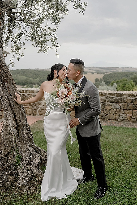 Wedding couple portrait of bride and groom kiss beside an olive tree, bride holding a rose bouquet in a strapless satin dress by a stone wall