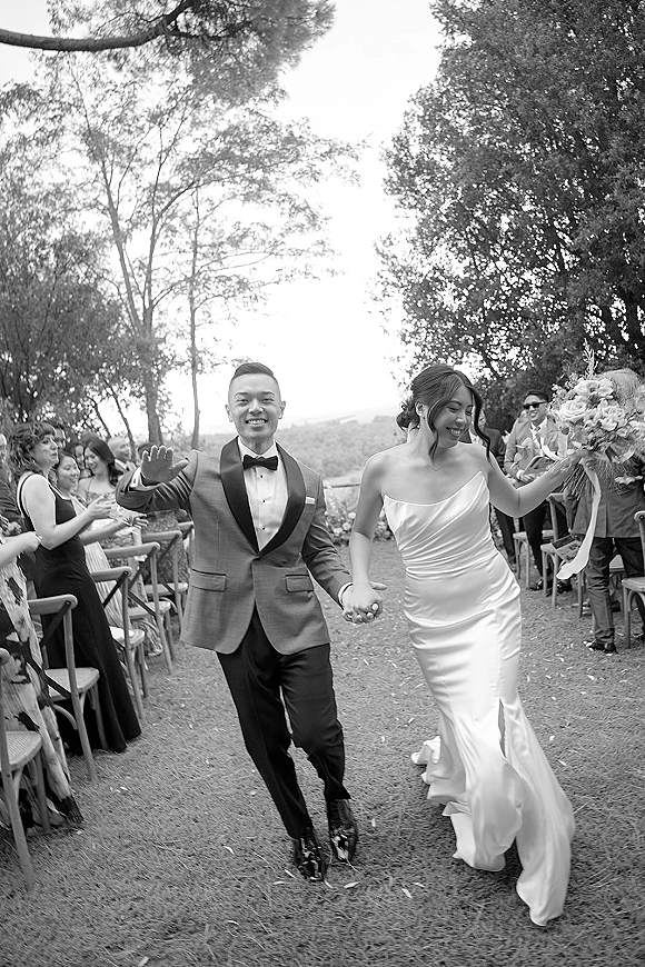 Wedding recessional as bride and groom walk hand in hand down the outdoor aisle, bride holding bouquet with ribbon, groom waving to guests
