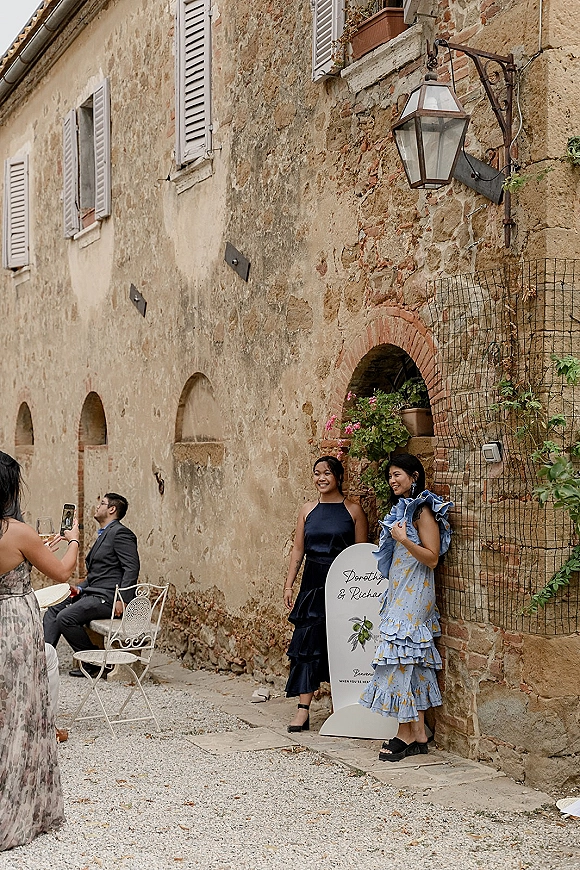 Wedding guest photo of three guests posing beside a wedding welcome sign, holding champagne, in a gravel courtyard with stone wall and arched doorway