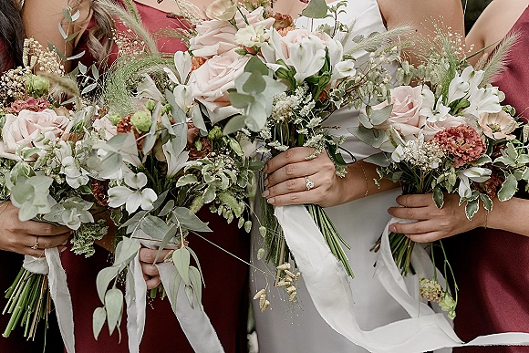 Bridesmaid bouquets of blush roses and white flowers with eucalyptus and ribbon streamers, held against a neutral backdrop with dresses and jewelry visible
