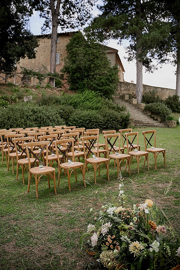Ceremony seating setup with crossback chairs lining a flower-marked aisle on a lawn, leading to stone steps beside a farmhouse and trees