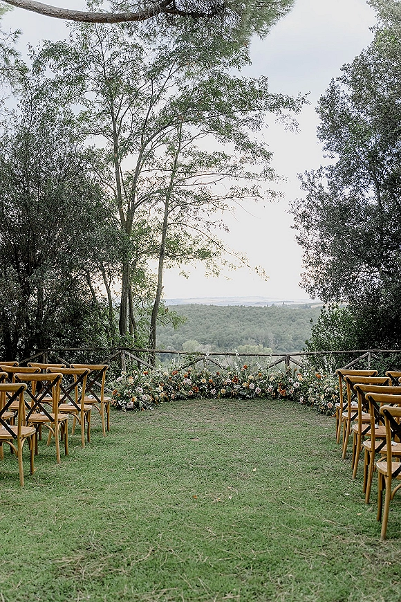 Ceremony setup for an outdoor wedding ceremony with wood chairs and floral ground arrangement on a grass lawn overlooking forest hills