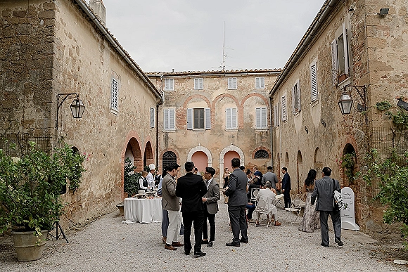 Cocktail hour with wedding cocktail hour guests mingling around white-clothed tables in a stone courtyard with arched doorways and lanterns