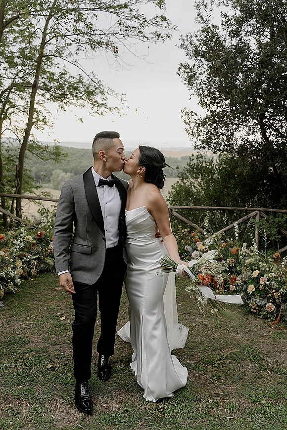 Wedding kiss portrait of bride and groom kissing, she in strapless satin dress holding bouquet, outdoors by wooden fence and hills