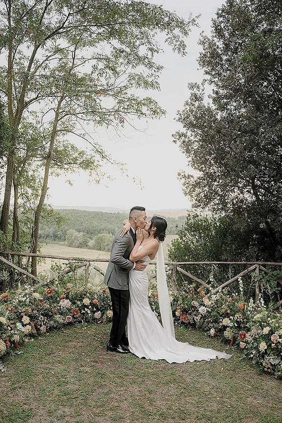 Wedding kiss as the bride and groom kissing, her long veil trailing over a floral ground arrangement by a wooden fence hillside backdrop
