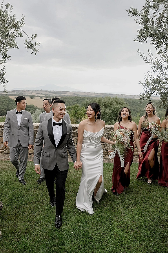 Wedding party portrait of bride and groom walking with bridesmaids and groomsmen, bride holding bouquet on grass lawn by stone wall