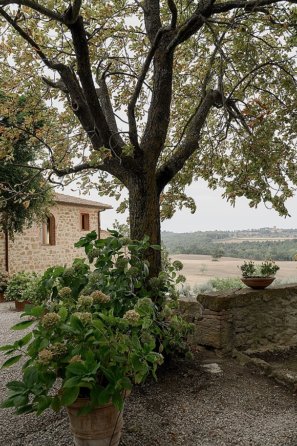 Outdoor wedding venue courtyard with potted plants and a stone planter along a gravel path beside a large tree and stone building under overcast sky