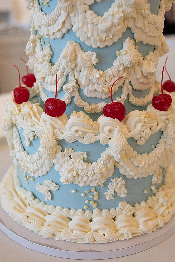 Wedding cake with blue wedding cake fondant, white buttercream piping swags, sugar pearls, and red maraschino cherries on a tabletop