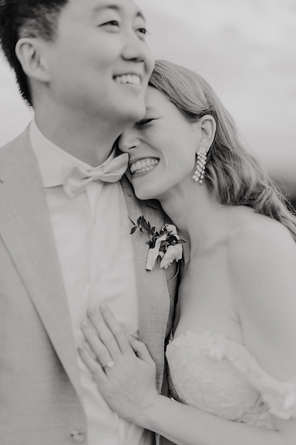 Couple portrait in a black and white wedding portrait, bride hugging groom, showing his boutonniere and her wedding ring against open sky