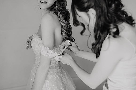 Getting ready moment as a bride getting dressed, showing lace bodice and off-the-shoulder sleeves while adjusting earrings by a plain wall