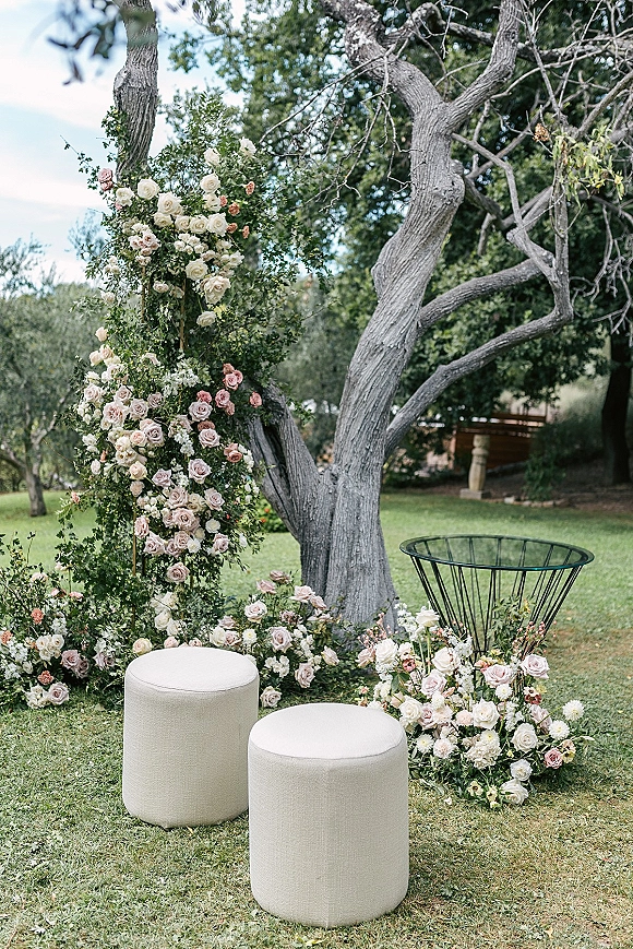 Ceremony floral installation with outdoor ceremony flowers on a wooden frame, roses and greenery with ivory poufs beneath a mature garden tree