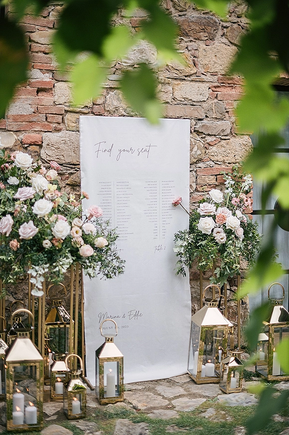 Wedding seating chart with calligraphy text on a sign, framed by blush and white roses with greenery on gold stands against a stone wall