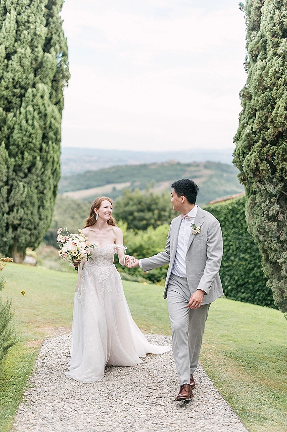 Couple portrait of bride and groom walking hand in hand, bride holding bouquet on a gravel path lined with cypress trees