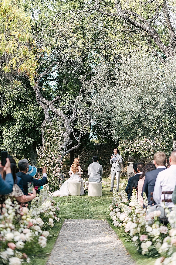 Wedding ceremony with outdoor wedding ceremony aisle flowers leading to a floral arch, bride and groom seated as officiant speaks in garden setting