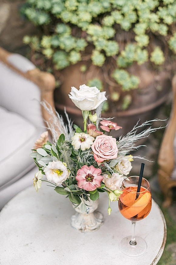 Wedding centerpiece with blush and white rose blooms in a vase beside an orange spritz cocktail on a round side table in an outdoor lounge area