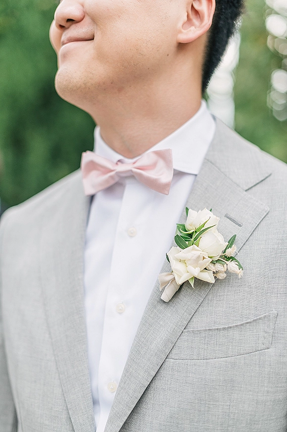 Groom boutonniere with a white rose boutonniere and greenery pinned to a light gray suit, paired with a light pink bow tie outdoors