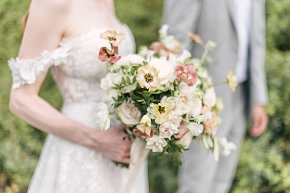 Bridal bouquet of garden rose bridal bouquet in blush and ivory with greenery and ribbon, held by bride in off-shoulder lace dress beside groom in light gray suit