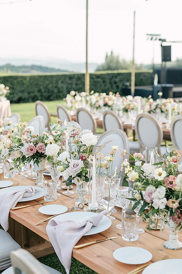Reception tablescape with an outdoor reception table on a wood farm table, featuring rose centerpieces, taper candles, and crystal glassware on a lawn