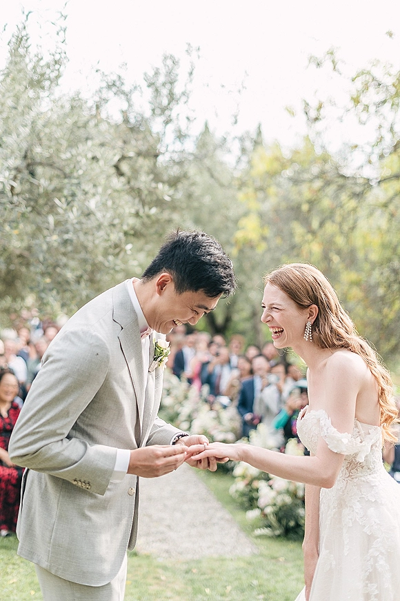 Ceremony moment during ring exchange as bride and groom laugh, he places wedding rings beside white aisle florals in a garden setting