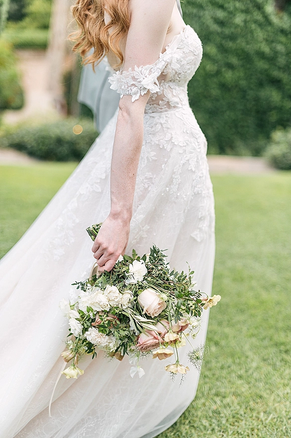 Bridal portrait of a bride holding bouquet of white and blush roses with greenery, wearing an off-the-shoulder lace gown on a garden path