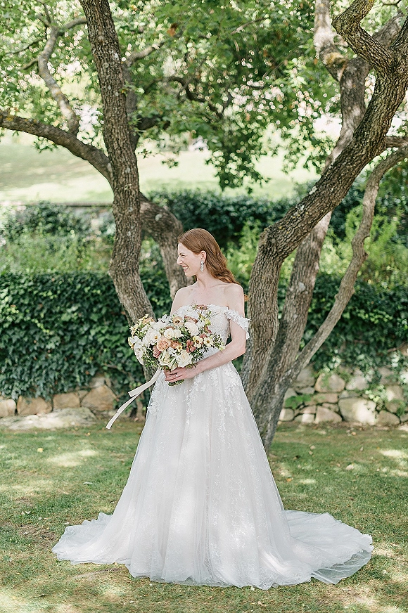 Bridal portrait of a bride holding bouquet in a strapless lace wedding dress with ribbon tie and drop earrings by a stone wall and hedge