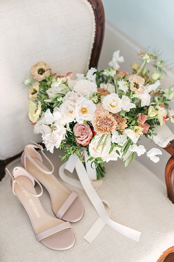 Bridal bouquet with pastel wedding bouquet blooms and a trailing white ribbon beside blush ankle strap heels on an upholstered chair