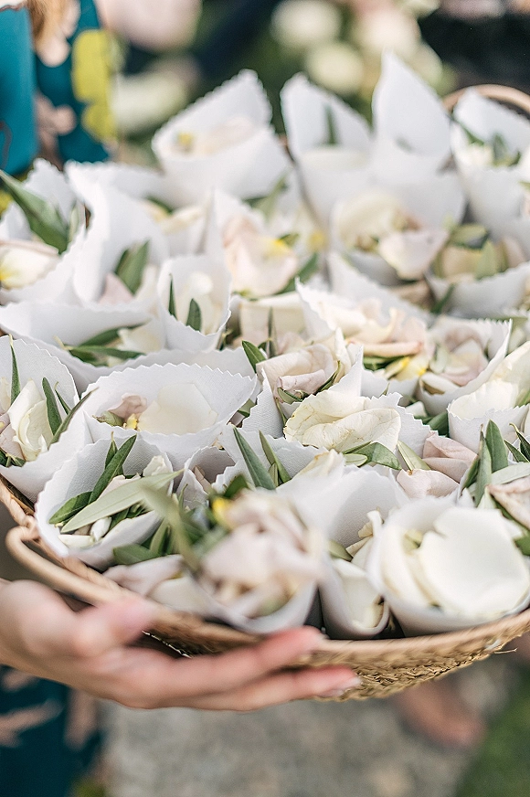 Flower petal confetti in paper cones sits in a woven basket, with white and blush rose petals held by hands amid blurred guests outdoors