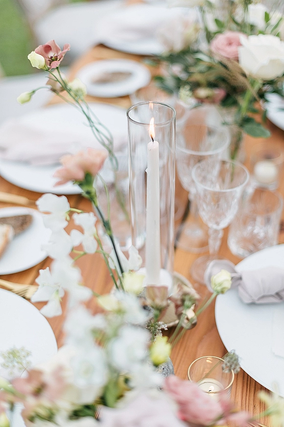 Reception tablescape with a wedding table centerpiece of taper candles in glass hurricanes, bud vases, and place settings on a wood table