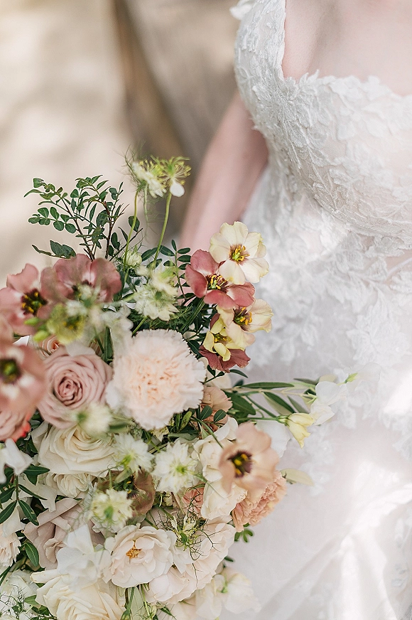 Bridal bouquet of blush and cream bouquet roses with greenery held against an ivory lace wedding dress in soft light by a neutral wall