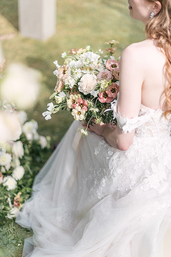 Bridal portrait of bride holding bouquet, wearing off-the-shoulder lace dress with tulle skirt and crystal earrings on a lawn by a white ceremony structure
