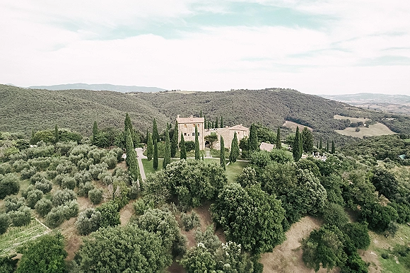 Wedding venue landscape with a stone villa and cypress trees lining a gravel driveway, set amid rolling hills and farmland under open sky