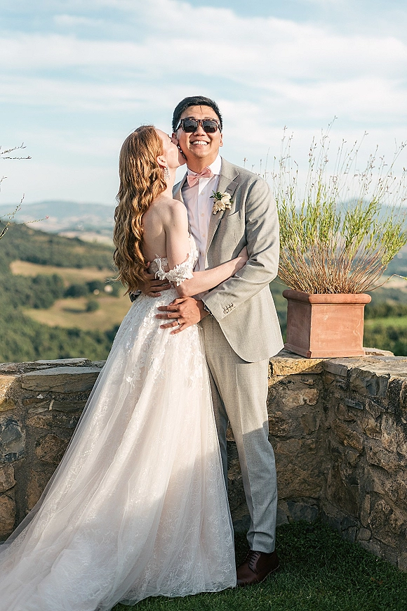 Couple portrait of bride kissing groom’s cheek, her lace off-shoulder dress and veil beside his sunglasses by a stone wall hillside view