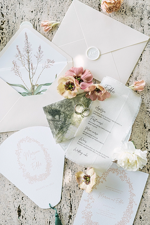 Wedding invitation suite wedding stationery flat lay with wax-sealed envelopes, floral cards, vellum ribbon, rings, tassel and flowers on stone surface