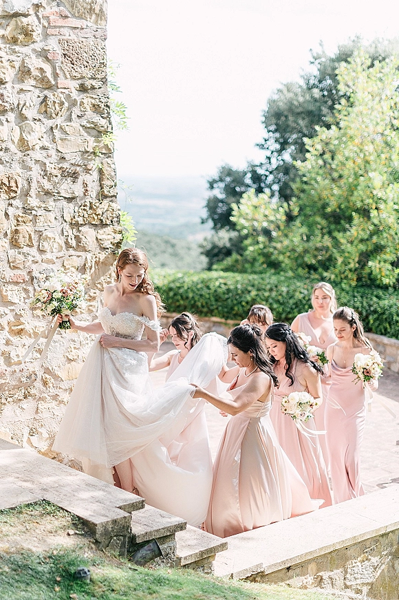 Bride with bridesmaids helping adjust her veil and train on stone steps, off-the-shoulder lace gown and blush dresses on a garden terrace