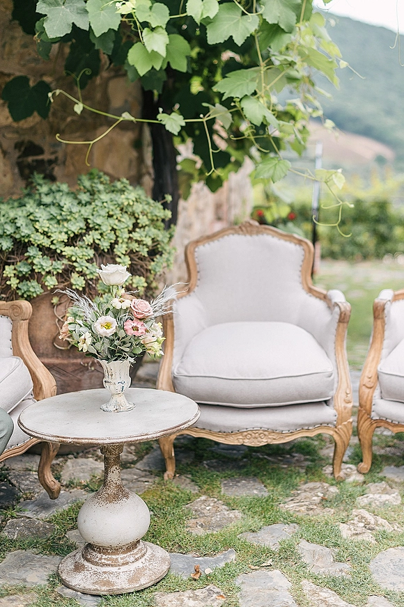 Lounge seating decor with cream upholstered armchairs and a pedestal table topped with a floral vase on a stone patio by a rustic wall