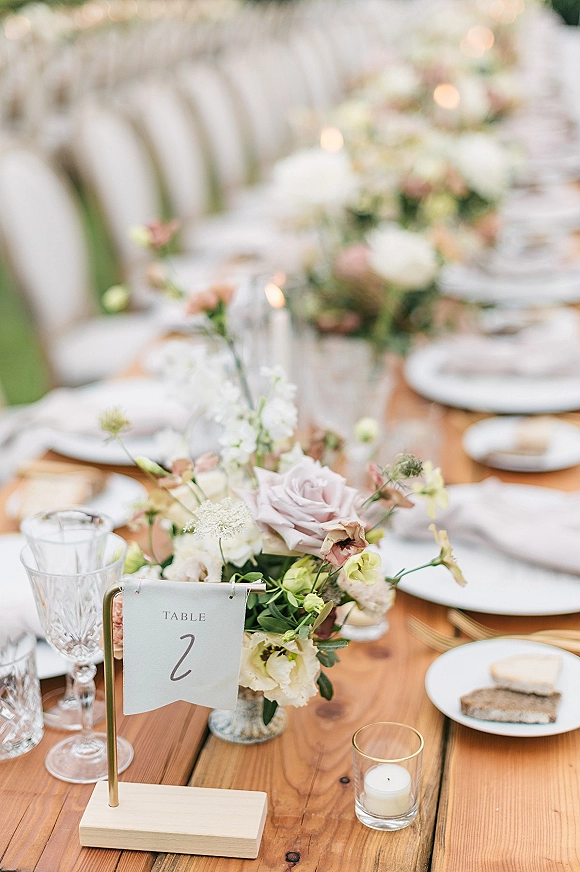Reception tablescape on a wood farm table wedding setup with rose centerpiece and taper candles, set with plates, goblets, and cake slices