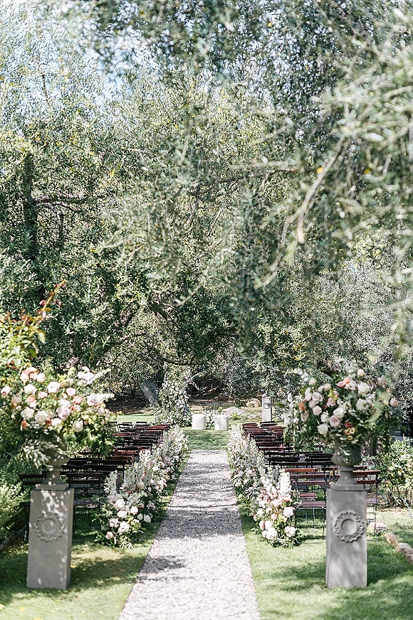 Ceremony aisle decor with outdoor wedding ceremony setup, pink and white florals in urns beside wooden chairs along a gravel runner on a garden lawn