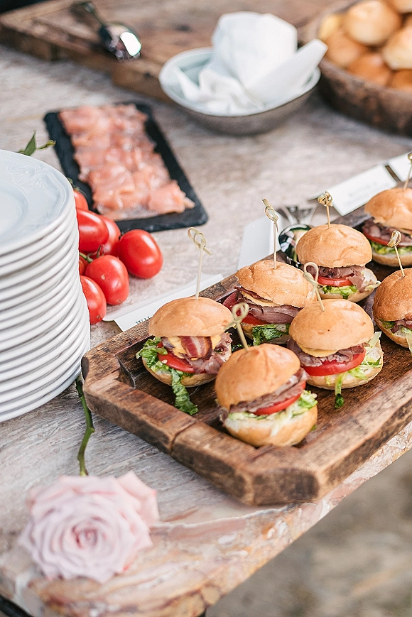 Wedding slider appetizers with mini burger sliders on a wooden tray with bamboo picks, cherry tomatoes and cheese on a rustic table, rose accent