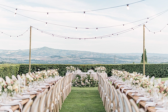 Outdoor reception setup with long banquet tables, string lights, and floral garlands on a lawn with hedge and mountain views