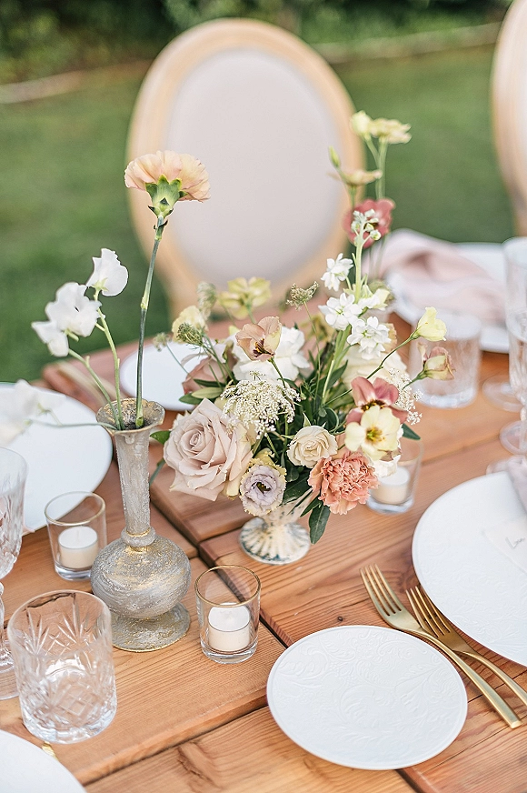 Reception tablescape with wildflower wedding centerpiece and bud vases, blush napkins, gold flatware, votive candles on a wooden table on a green lawn