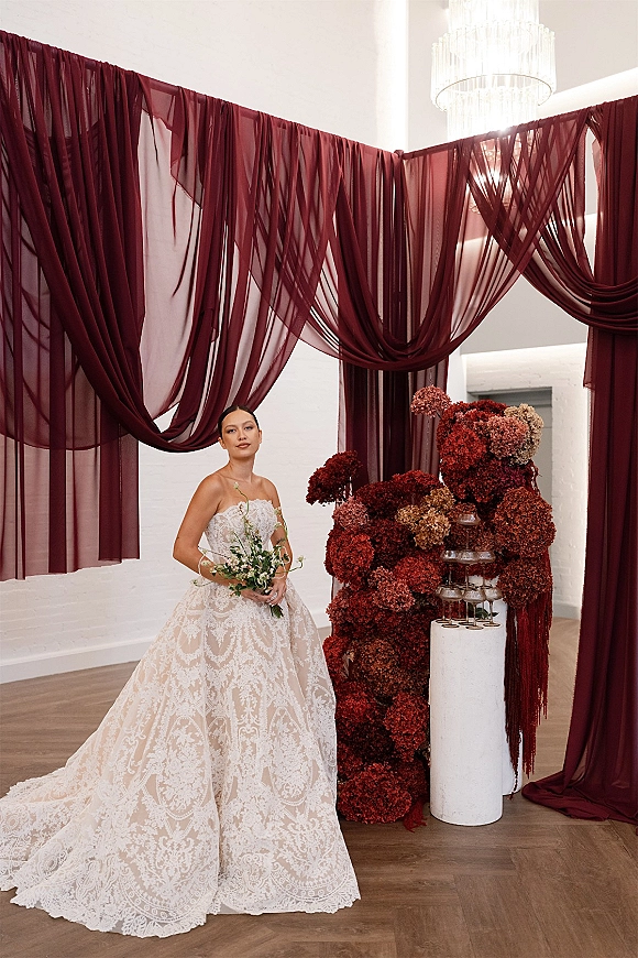 Bridal portrait in a strapless lace wedding dress, holding a greenery bouquet beside a champagne tower under a chandelier in a white-walled venue