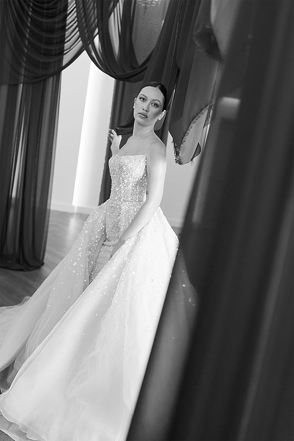 Bridal portrait in black and white of a bride in a strapless beaded bodice and tulle skirt with long train, posed by window and curtains