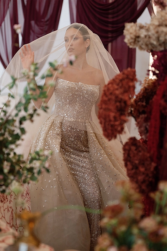 Bridal portrait of a bride with veil over her face in a strapless beaded gown with tulle overskirt, framed by burgundy drapery and florals