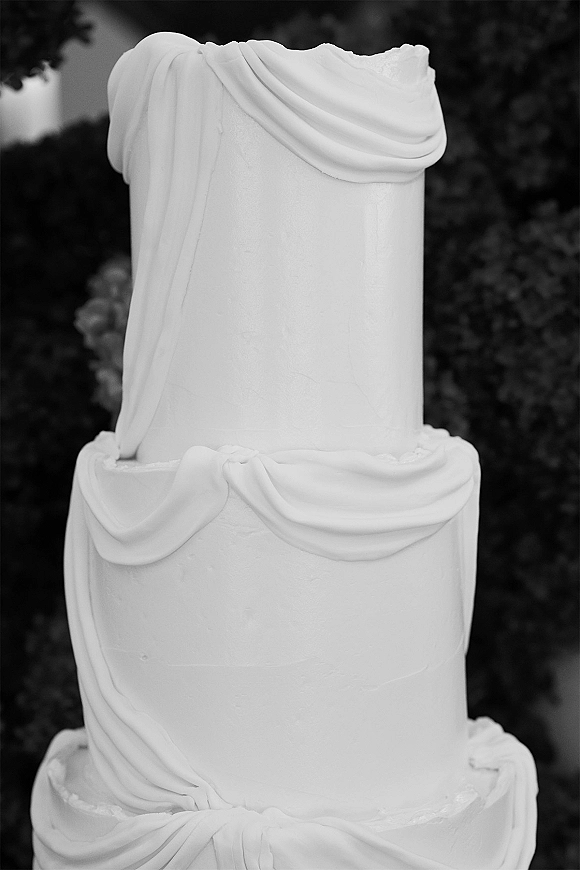 Wedding cake with white wedding cake fondant draping on a three-tier design, set against softly blurred outdoor greenery background