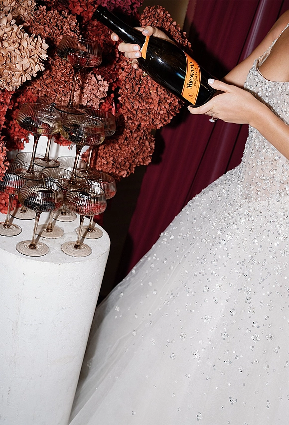 Champagne tower of stacked coupe glasses as a sequin wedding dress hand with engagement ring pours from a bottle against burgundy drapery