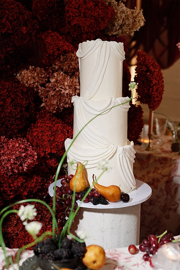 Wedding cake with draped fondant frosting on a stand, styled with pears, grapes, and berries beneath a dark red floral installation