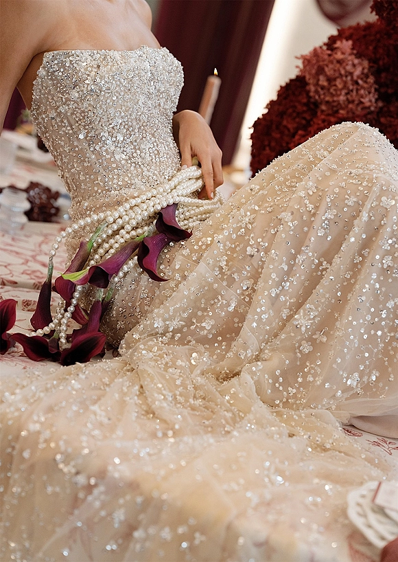 Beaded wedding dress and strapless bridal gown laid on a table with crystal sequins, pearl strands, calla lily bouquet, and a candle before burgundy drapery