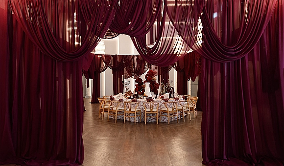 Reception tablescape with burgundy wedding draping over a round table set with floral centerpieces, taper candles, and a white wedding cake indoors