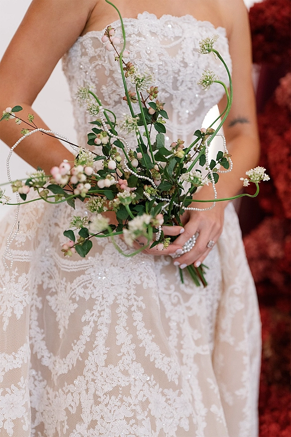 Bridal bouquet wildflower bridal bouquet with white blooms, greenery, pink berries and pearl strands held against a strapless lace bodice by red wall