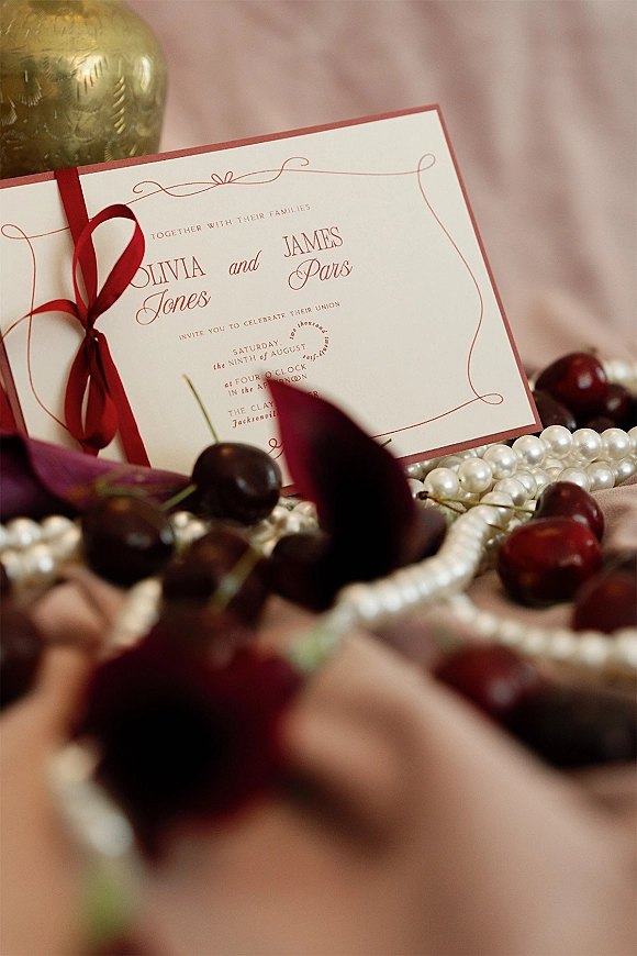 Wedding invitation suite with red wedding invitations tied in a red ribbon, styled with pearls and cherries on pink fabric with petals